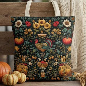 Op de afbeelding: Een zwarte draagtas met een kleurrijk bloemen- en kalkoenontwerp. De tas heeft zonnebloemen, pompoenen en een kalkoen in het midden. De tas heeft crèmekleurige handvatten en staat op een houten bank.