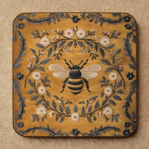 May include: Square wooden coaster with a yellow background and a detailed bee illustration. The bee is surrounded by a floral wreath with white flowers and gray leaves. The coaster has a dark brown border.