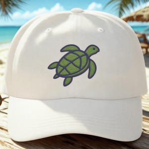 May include: A white baseball cap with a green and blue embroidered sea turtle design. The cap is on a wooden surface with a beach and ocean background. The cap has a curved brim and a rounded crown.