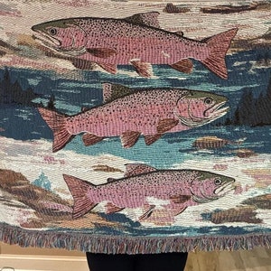 May include: A woven tapestry throw blanket featuring three pink trout swimming in a blue river. The background is a light brown and white with a dark green forest on the right side of the image.
