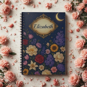 May include: A spiral-bound notebook with a navy blue cover featuring a floral design and the name "Elizabeth" in a gold frame. The design includes various flowers, a crescent moon, and stars.