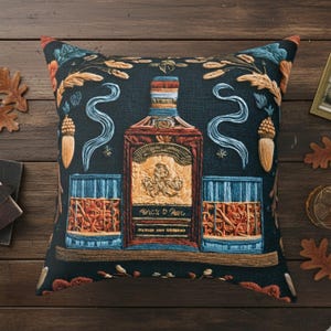 May include: Decorative square pillow with a dark background featuring a whiskey bottle and two glasses. The bottle has a detailed label and is flanked by stylized swirls and acorns. The pillow has a rustic, vintage aesthetic.