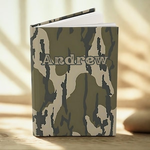 May include: A personalized notebook with the name "Andrew" printed on the cover. The cover features a camouflage pattern in shades of green, brown, and beige. The notebook is open, revealing white pages.
