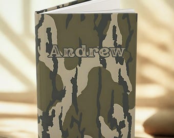 Puede incluir: Un cuaderno personalizado con el nombre "Andrew" impreso en la portada. La portada presenta un patrón de camuflaje en tonos verdes, marrones y beige. El cuaderno está abierto, revelando páginas blancas.