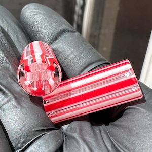 Peut inclure: Deux composants cylindriques en verre présentant un motif rayé de verre rouge, blanc et transparent alterné. Une pièce est positionnée verticalement pour montrer la structure interne de six canaux circulaires, tandis que l'autre est posée horizontalement.