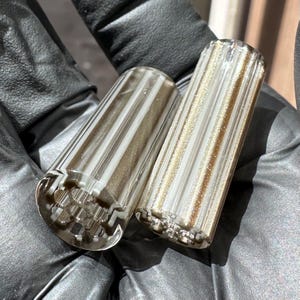 May include: Two cylindrical glass beads featuring vertical stripes of opaque white and translucent gold glitter. The beads have a clear glass exterior with a multi-faceted interior core visible at the ends, creating a textured, reflective appearance.