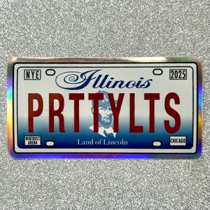 May include: A blue and white Illinois license plate with the text "PRTTYLTS" in red letters. The plate has the text "Nye" and "2025" in the top corners. The plate also has the text "Wintrust Arena" and "Land of Lincoln" in the bottom corners.