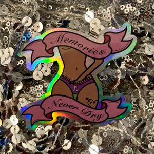 May include: A holographic sticker with a pink ribbon that says "Memories Never Dry". The sticker features a cartoon illustration of a person with brown skin wearing underwear and a tattoo of an arrow and heart.
