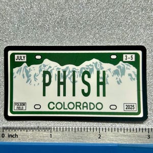 May include: A rectangular sticker resembling a Colorado license plate. The sticker is green and white, with the word "PHISH" in large green letters. It includes the text "COLORADO" and "2025", and the text "JULY" and "3-5".
