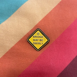 Op de afbeelding: Een geel, ruitvormig waarschuwingsbord met een zwarte rand en zwarte tekst. Het bord luidt "BRIDGE MAY BE SWIRLY" met een spiraalvormig symbool boven het woord "BRIDGE".