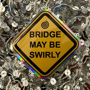 Może przedstawiać: Żółty znak ostrzegawczy w kształcie rombu z czarną obwódką i symbolem spirali. Znak informuje "Bridge May Be Swirly".