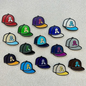 Op de afbeelding: Een verzameling van 15 emaille-pins in de vorm van baseballpetten in verschillende kleuren, elk met een gestileerd letter "R"-logo.