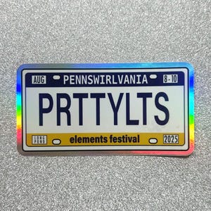 May include: A rectangular sticker with a holographic border, resembling a license plate. The sticker reads "PRTTYLYTS" in large blue letters. Above, it says "PENNSWIRLVANIA" and "AUG 8-10". Below, it says "elements festival" and "2025".