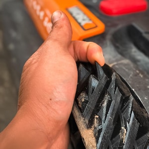 May include: Close-up of a black rubber sole with a deep tread pattern. The sole is held by a hand, with an orange object and a red object in the background. The tread is designed for grip and traction.