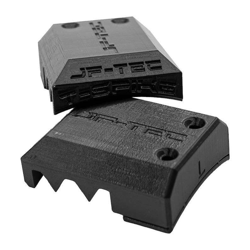 May include: Two black, angular, plastic components with the text "JF-TEC" embossed on the surface. One component is positioned on top of the other. The bottom component has a series of triangular teeth on one edge and the letter "L" on the side.