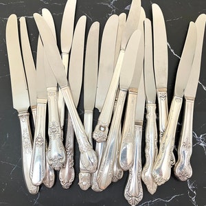May include: A collection of vintage silver butter knives with ornate handles. The knives are arranged in a fan shape, with the handles facing the viewer.
