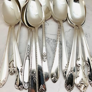 May include: A collection of polished silver spoons with ornate handle designs. The spoons have a reflective surface, and the handles feature various floral and geometric patterns. The spoons are arranged on a light-colored surface.