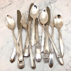 May include: A collection of assorted silver-toned flatware including several spoons, forks, and knives. The pieces feature various handle designs, ranging from smooth, plain surfaces to intricate floral and geometric embossed patterns.