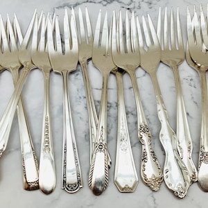 May include: A collection of 13 silver forks with ornate handles. The forks are arranged in a fan shape on a white marble surface.