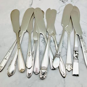 May include: A collection of vintage silver butter knives with various handle designs. The knives are arranged on a white and gray marble surface, showcasing their polished, reflective surfaces. The blades are flat and wide.
