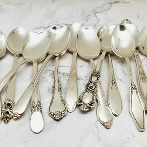 May include: A collection of vintage silver spoons with ornate designs. The spoons are arranged in a fan shape on a white marble surface.