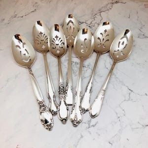 May include: A collection of silver serving spoons with ornate handles and slotted bowls. The spoons are arranged on a marble surface, showcasing their intricate floral and scrollwork designs. The spoons are made of a shiny, reflective metal.