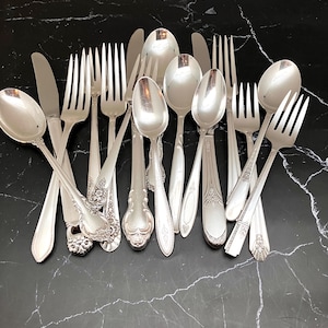 May include: An assortment of vintage silverware, including forks, spoons, and knives, arranged on a black marble surface. The silverware has a shiny, silver finish, with various decorative handle designs. The utensils are of different sizes and shapes.