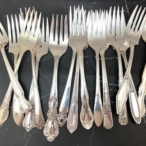 May include: A collection of 15 silver forks with ornate handles. The forks are arranged in a fan shape, with the handles pointing towards the top of the image.