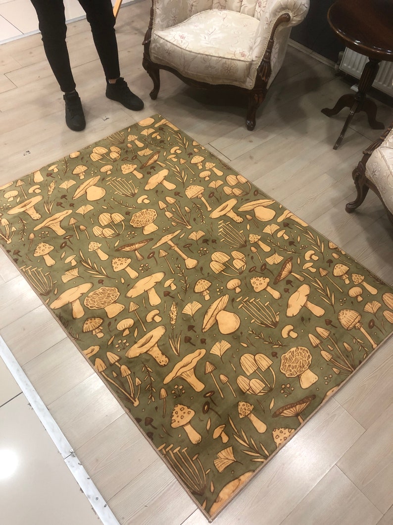 May include: A green and beige area rug with a mushroom pattern. The rug has a beige border and features a variety of mushroom shapes and sizes.