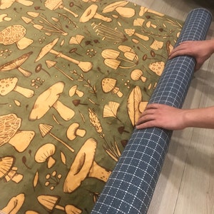 May include: A green rug with a repeating pattern of brown mushrooms and leaves. The rug is rolled up and being held by a hand.