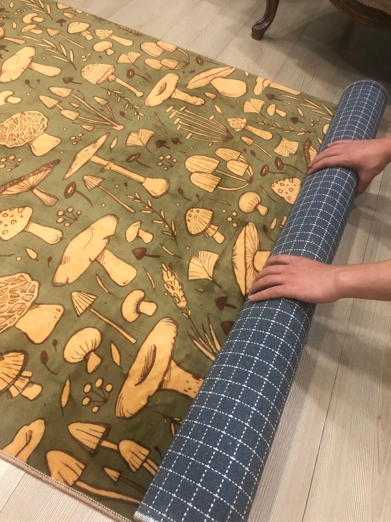 May include: A green rug with a repeating pattern of brown mushrooms and leaves. The rug is rolled up and being held by a hand.
