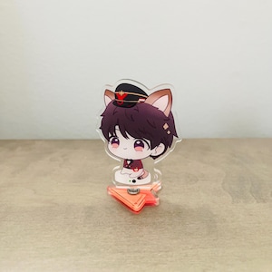May include: A clear acrylic standee featuring a chibi-style character with dark hair, cat ears, and a military-style cap. The character wears a dark red jacket with a black bowtie and sits on a translucent orange, arrow-shaped base.