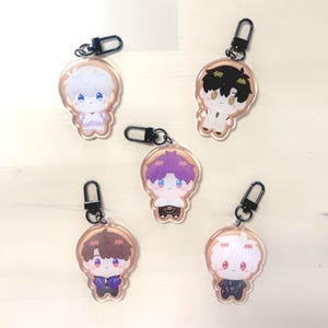 Op de afbeelding: Vijf acryl sleutelhangers met chibi-achtige anime-personages met verschillende haarkleuren en outfits. Elke sleutelhanger heeft een zwarte sluiting. De personages hebben grote ogen en bevinden zich in verschillende poses. De sleutelhangers liggen op een lichtgekleurd oppervlak.