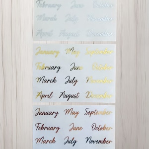 May include: Three sheets of white paper with the months of the year written in different colors. The first sheet has silver lettering, the second sheet has gold lettering, and the third sheet has copper and blue lettering.