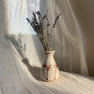 Op de afbeelding: Een witte keramische vaas met een bruine glazuur, met een druppelontwerp, bevat een boeket gedroogde lavendelbloemen.