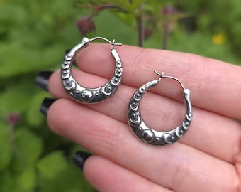 May include: A pair of silver hoop earrings. Each earring features a crescent shape with a series of small, raised circular and crescent-shaped designs. The earrings are held against a blurred green background.