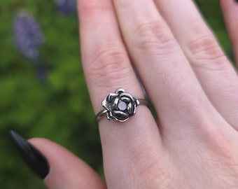 Könnte beinhalten: Ein silberner Ring mit einem detaillierten Rosenmuster. Der Ring wird an einem Finger getragen, wodurch das komplizierte Blumenmuster hervorgehoben wird. Der Hintergrund ist verschwommen, mit Andeutungen von Grün und Lila.