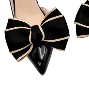 May include: A pair of black patent leather pumps with a pointed toe and a large black bow with cream-colored trim. The shoes have a low heel and a cutout design on the side.