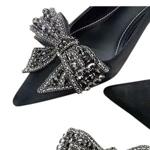 May include: Black pointed-toe pumps with a decorative bow made of black and silver rhinestones. The bow is a prominent feature, adding a touch of glamour to the footwear. The image shows the shoe from multiple angles.