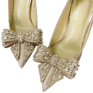 May include: A pair of champagne-colored high heels with pointed toes. Each shoe features a large, embellished bow with sparkling crystals. The heels have a metallic sheen, and the inner soles are a light gold color.