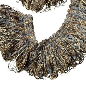 May include: A close-up of a gold, brown, and grey tassel trim. The trim is made of a soft, fluffy material and has a fringe-like appearance.