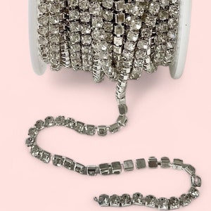 May include: A spool of silver rhinestone chain. The chain is made of small, square-cut rhinestones linked together. The chain is unspooled from a white plastic spool, with a portion of the chain lying flat on a pink background.