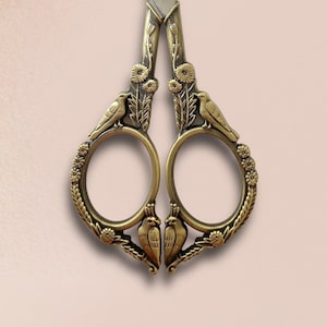 May include: Vintage-style embroidery scissors with a decorative bronze finish. The scissors feature a detailed design with birds and floral elements. The blades are silver-colored, and the handles have a decorative, ornate pattern. These scissors are ideal for sewing and crafting.