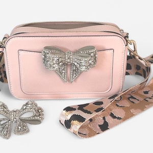 May include: A pink camera bag with a large silver bow detail and a leopard print strap.