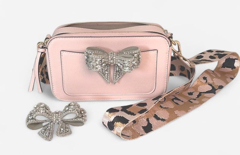 May include: A pink camera bag with a large silver bow detail and a leopard print strap.