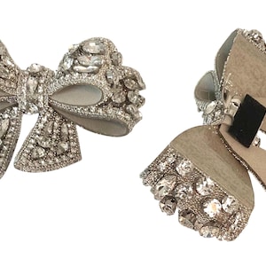 May include: Two silver bow-shaped brooches with rhinestones. One bow is made of fabric and the other is made of leather.