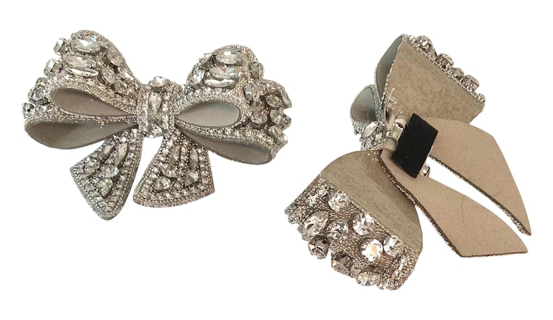 May include: Two silver bow-shaped brooches with rhinestones. One bow is made of fabric and the other is made of leather.