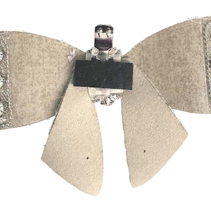 May include: A beige fabric bow with a silver metal clip and clear rhinestones. The bow is decorated with a silver glitter finish.
