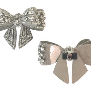 May include: Two silver bow brooches with rhinestones. The top brooch has a more traditional bow shape, while the bottom brooch has a more modern, flat bow shape.