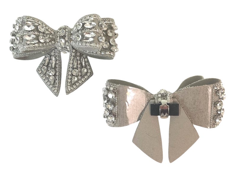 May include: Two silver bow brooches with rhinestones. The top brooch has a more traditional bow shape, while the bottom brooch has a more modern, flat bow shape.
